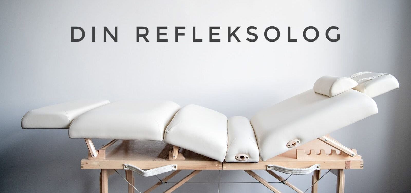 Behandlingsbenk i lyst tre og hvit polstring står mot en lys vegg med teksten "Din Refleksolog".